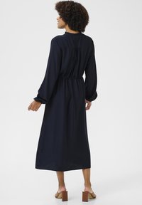 Námornícky modré šaty s dlhým rukávom, frilovaným výstrihom, zhromaždeným dizajnom na chrbte a s úzkym pásom. Modelka má na sebe hnedé sandále na blokovom podpätku.