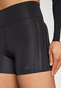 Pantaloni sportivi neri con vita alta, tessuto morbido, strisce diagonali scure a contrasto sui lati e un piccolo logo vicino all’orlo.