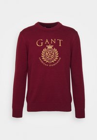 Pull en maille bordeaux avec un col rond, des manches longues et un logo doré orné d'éléments décoratifs au centre.