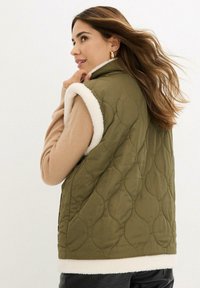 Gilet trapuntato verde oliva con colletto in pile crema, caratterizzato da una vestibilità ampia. È indossato sopra un maglione beige, mostrando texture morbide e sovrapposizioni.