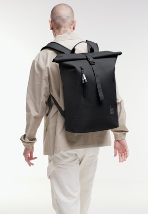 Persoon draagt beige jas en broek, draagt een grote zwarte roll-top rugzak met voorrits en "GOT BAG" logo op de achterkant.