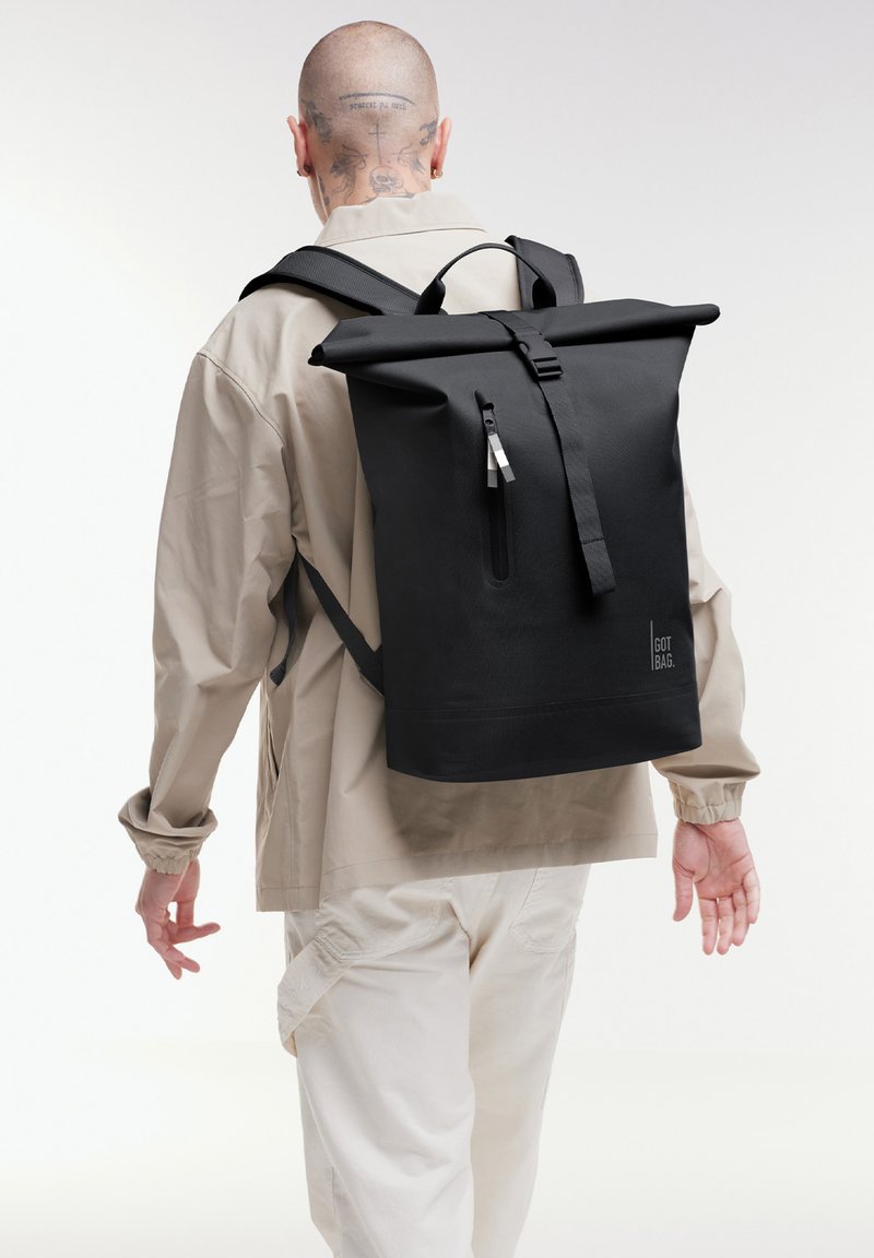 Personne portant une veste et un pantalon beige, avec un grand sac à dos noir à rabat enroulable, une fermeture éclair à l'avant et le logo "GOT BAG" au dos.