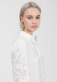 Camicia bianca con bottoni, decorata con ricami floreali dettagliati su una manica, presenta perline e paillettes, texture liscia e colletto classico.