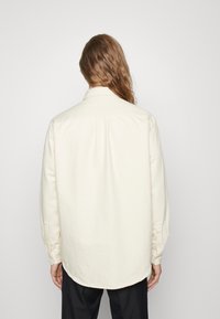 Camicia a maniche lunghe color crema con colletto classico, orlo curvo e polsini con bottone, realizzata in un tessuto morbido. Design semplice e minimalista.