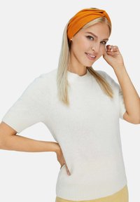 Maglione bianco a maniche corte con scollo rotondo e polsini a coste. Fascetta per capelli arancione e texture. Capelli chiari. In piedi con una mano sulla vita.