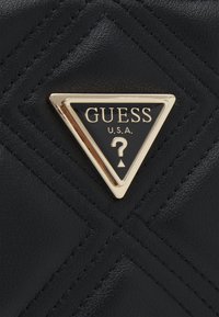 Surface en cuir noir matelassé avec un emblème triangulaire bordé d’or portant l’inscription « GUESS U.S.A. » et un point d’interrogation au centre.