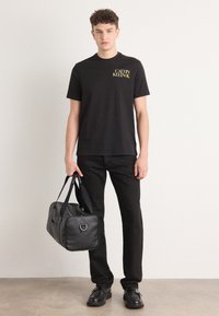 Sort bomulds T-shirt med gult "Calvin Klein" logo, parret med sorte bukser og en sort læder duffel taske med en sølv hardware accent.