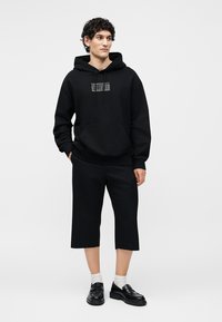 Jeune homme portant un sweat à capuche noir avec poche avant, pantalon court noir, chaussettes blanches et mocassins noirs, debout les mains détendues.