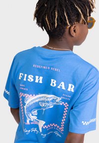 Camiseta de algodón azul con un gran gráfico de pez y texto. Mangas cortas, corte relajado, con detalles a cuadros en rosa y blanco.