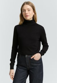 Mustrasmudel must turtleneck-kampsun, vöödistruktuuriga, sobiv disain pikkade varrukatega. Kombineeritud tumedate teksadega, käed taskutes.
