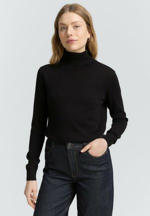 Maglione nero a collo alto in materiale a costine, design aderente con maniche lunghe. Abbinato a jeans in denim scuro, mani nelle tasche.