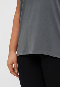 Grey, sleeveless top made of soft fabric, featuring a rounded hem and a relaxed fit, paired with black leggings.