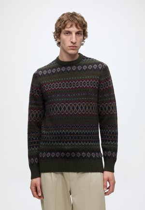 Jeune homme aux cheveux courts et bouclés portant un pull vert foncé à motif et un pantalon beige, debout devant un fond blanc uni.