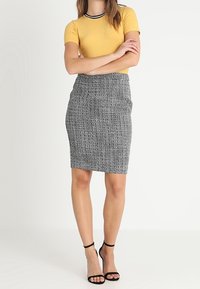 Gelbes, geripptes Kurzarmoberteil mit schwarzen Akzenten, kombiniert mit einem knielangen, schwarz-weißen gemusterten Bleistiftrock und schwarzen Riemchenheels.