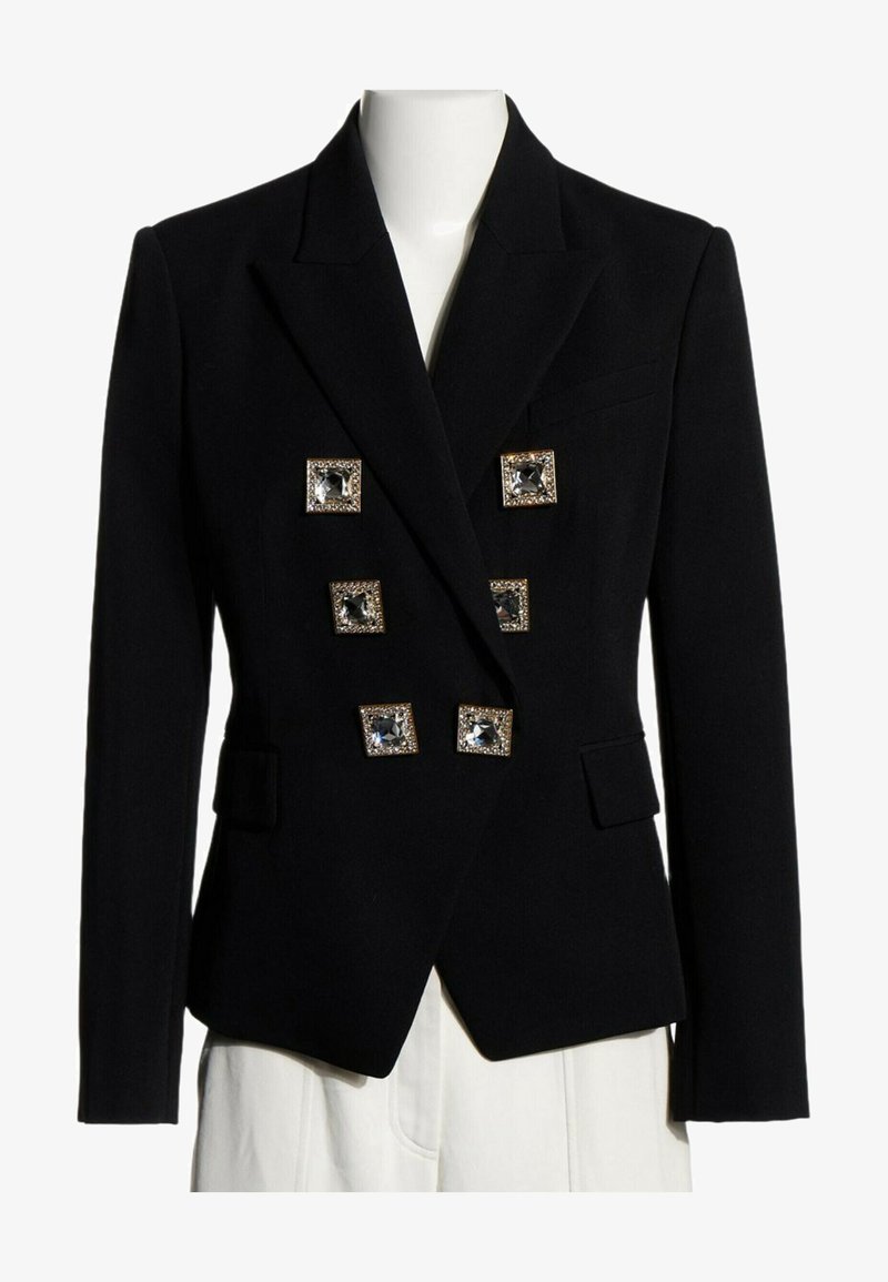 Blazer noir tailleur croisé avec six grands boutons carrés ornés de bijoux et poches à rabats, présenté sur un mannequin portant un pantalon blanc.