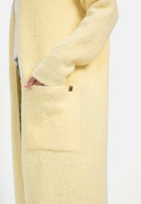 Hoge, zachte cardigan van gestructureerde, lichtgele stof met een voorke pocket en subtiele stikdetails langs de randen.