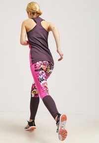 Mujer corriendo en interiores, vestida con una camiseta sin mangas de color púrpura y leggins florales en rosa y negro, con zapatillas de correr en negro y rosa.