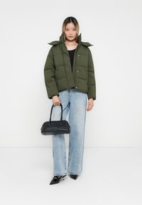 Chaqueta acolchada verde oscuro con cierres a presión, bolso negro, jeans de pierna ancha azul claro y tacones de punta negra.