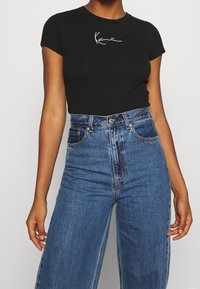 Černé přiléhavé tričko s krátkými rukávy, se vzorem bílého ručně psaného loga. Kombinováno s vysokými světle modrými džínovými kalhotami s širokými nohavicemi.