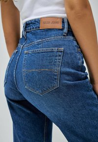 Modré denimové džíny s vysokým pasem, výraznými zadními kapsami a koženou nášivkou s nápisem "SALSA JEANS" na pasu.