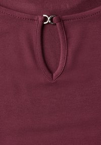 Burgundy topp med en strukturerad yta, med en nyckelhålöppning vid halsen och en silverdetalj vid stängningen.