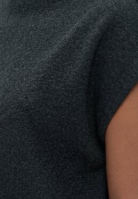 Gros plan sur un tissu texturé gris foncé couvrant l’épaule et le haut du bras d’une personne à la peau foncée.