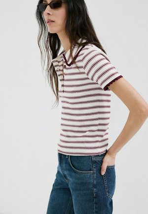 Kobieta w krótkim koszulce polo w paski i niebieskich dżinsach, w okularach przeciwsłonecznych, stoi z jedną ręką w tylnej kieszeni na jednolitym tle.