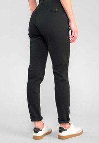 Zwarte slim-fit broek met een hoge taille, voorzien van een achterzak en opgerolde broekspijpen, gecombineerd met zwart-witte sneakers tegen een lichte achtergrond.