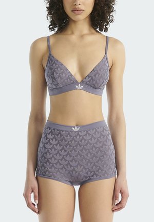 Woman wearing matching lavender bralette and high-waisted shorts with an Adidas trefoil pattern and logo on front bands.