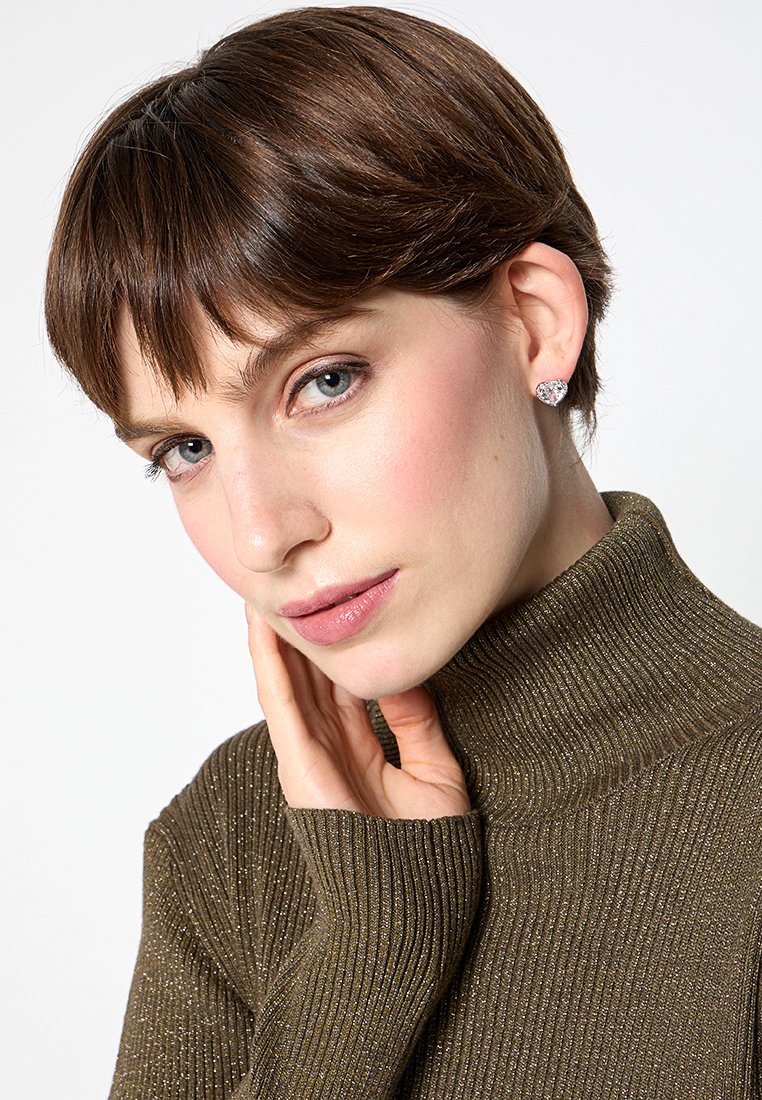 Model w ciemnozielonym, prążkowanym swetrze z golfem. Ma krótkie brązowe włosy i subtelny blask. Lekki makijaż z kolczykami w kształcie sztyftów.