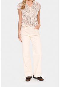 Blusa color beige chiaro con balze e pois abbinata a pantaloni a gamba larga color crema. Modello con mocassini marroni con dettagli decorativi.