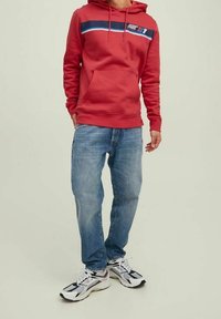 Hombre con sudadera roja con bolsillo frontal, jeans azules y zapatillas deportivas blancas, de pie contra un fondo claro y liso.