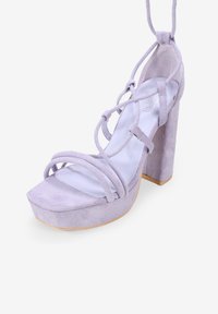 Sandalia de tacón alto de ante morado con tiras entrelazadas, punta cuadrada y un tacón grueso con superficie texturizada.