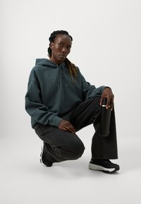 Felpa verde scuro con cappuccio e cordini, pantaloni neri a gamba larga e sneakers nere con dettagli bianchi. Tiene in mano una bottiglia d'acqua nera.