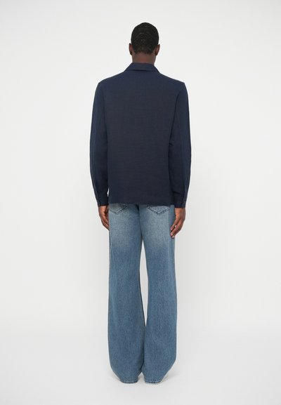 Moški stoji s hrbtom obrnjenim proti gledalcu, oblečen v temno modro srajco z dolgimi rokavi in široke svetlo modre kavbojke, v ozadju pa je enobarvna podlaga.