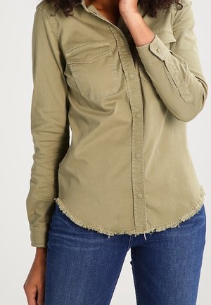 Kobieta w oliwkowo zielonej, długim rękawem, zapinanej na guziki koszuli z postrzępionym dołem i niebieskich dżinsach, ręka delikatnie dotyka twarzy.