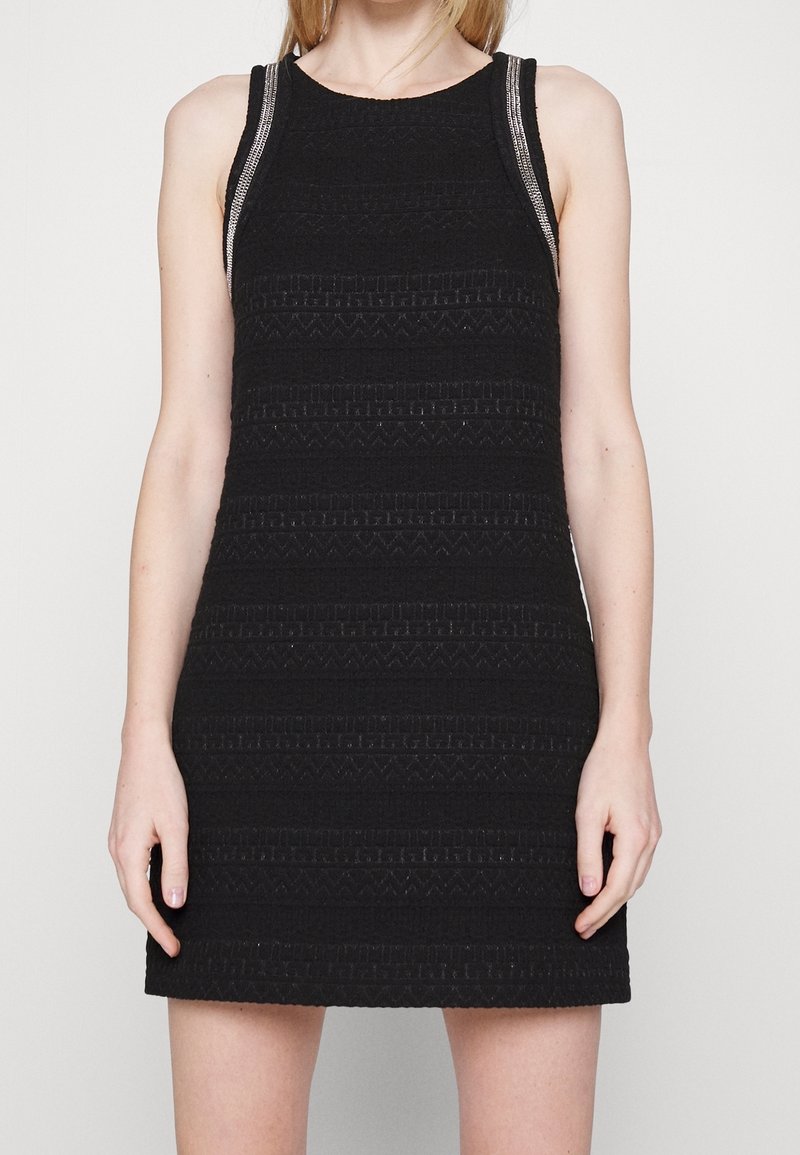Femme portant une mini robe noire texturée sans manches avec un galon argenté le long des bretelles, montrée du cou jusqu'à mi-cuisse.