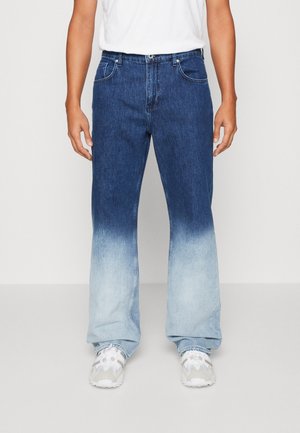 Mann trägt blaue Jeans mit einem hellblauen, verblassten Dip-Dye-Effekt von der Mitte des Oberschenkels bis zu den Säumen, kombiniert mit weißen Turnschuhen und einem weißen T-Shirt.