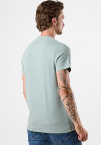 Ljusgrön t-shirt med korta ärmar, ribbad struktur och rund halsringning, som visar en figurnära design bakifrån. Diskreta sömdetaljer.