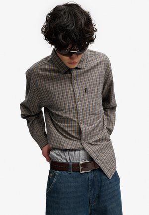Ung mand med krøllet mørkt hår iført en ternet skjorte, solbriller, bælte og jeans, der ser nedad med hænderne bag ryggen.