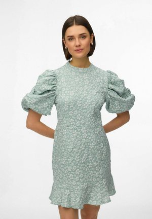 Rövid barna hajú nő világoskék virágos ruhában, dudorított rövid ujjakkal és fodros szegéllyel, kezét a háta mögött tartva áll.