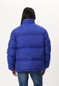 Chaqueta acolchada azul con un alto cuello, múltiples secciones acolchadas y puños elásticos. La tela parece suave con un acabado brillante.