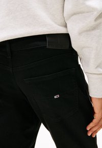 Osoba ubrana w czarne dżinsy z małym logo na tylnej kieszeni oraz jasnoszarą, długorękawową koszulę widoczną na wysokości talii i ramienia.