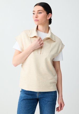 Femme aux cheveux foncés attachés en chignon, portant un haut beige sans manches zippé par-dessus un t-shirt blanc à manches courtes et un jean bleu, regardant vers la gauche.