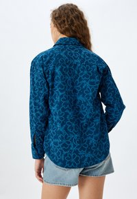 Blå skjorta med blommönster, långa ärmar och klassisk krage, med en lätt rundad nederkant och knappproppar på ärmsluten.