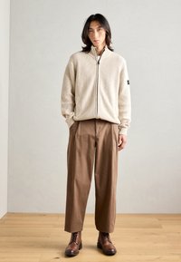 Krémový žebrovaný svetr na zip, hnědé široké kalhoty a hnědé šněrovací boty. Jednoduchý design s texturovanou tkaninou a neutrálními barvami.