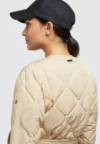 Beige doorgestikte pufferjas met een gladde textuur, voorzien van een riem en een zwart accentlabel aan de achterkant, gedragen met een zwarte pet.