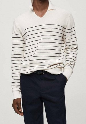 Homme portant un pull en maille blanc à fines rayures horizontales noires et un pantalon bleu marine foncé, une main dans la poche.