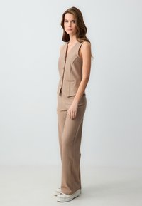 Kameez van tan kleur met pinstripe, een diepe V-hals en enkele knoopsluiting, gecombineerd met bijpassende loszittende broeken en witte sneakers.