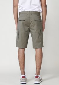 Uomo che indossa pantaloni cargo verde oliva, una maglietta bianca e scarpe da ginnastica bianche, visto da dietro su uno sfondo neutro.